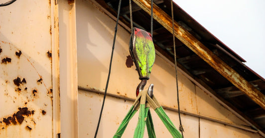 An old overhead crane hook lifting a heavy load with slings inside an industrial space with steel beams.