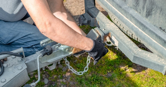 A close-up of a person fastening safety chains while connecting a trailer hitch to the rear of a vehicle.