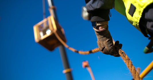 A low-angle view of a person at a construction site, holding a long rope attached to a box being lifted by a crane.